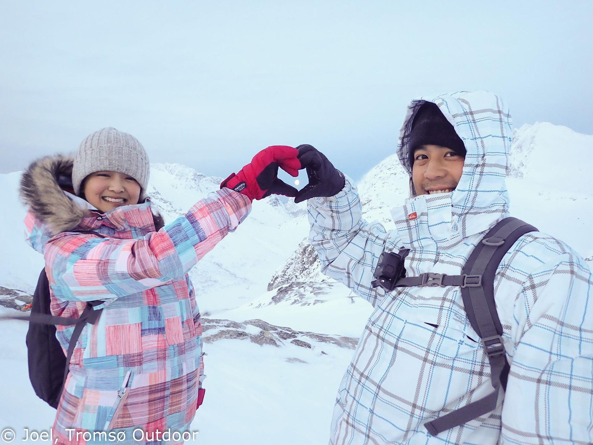 To personer i vinterklær lager hjertesymbol med hendene i snødekte fjell.