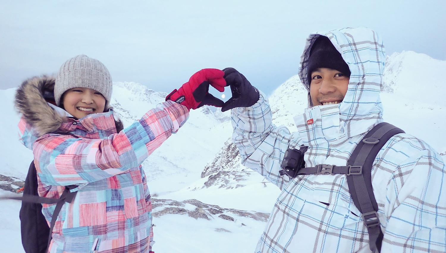Two people in winter clothing forming a heart shape with their hands in snowy mountains.