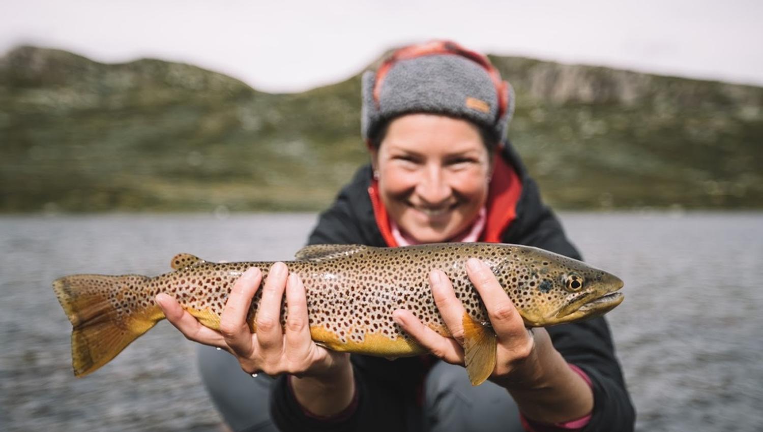Hilde from Vinternatt is holding a trout with Mountain in the background.