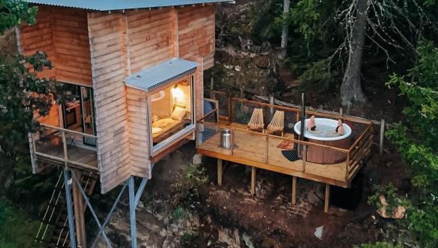 Small wooden cabin on stilts surrounded by forest, with landscape visible in the background.