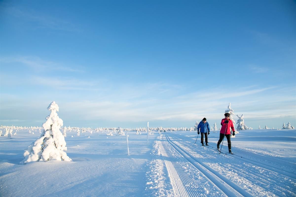 To skiløpere går i nypreparerte skispor over et åpent, snødekt fjellområde under klar blå himmel.