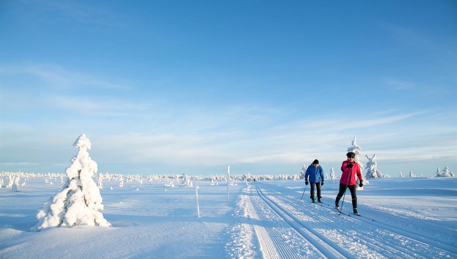 To skiløpere går i nypreparerte skispor over et åpent, snødekt fjellområde under klar blå himmel.