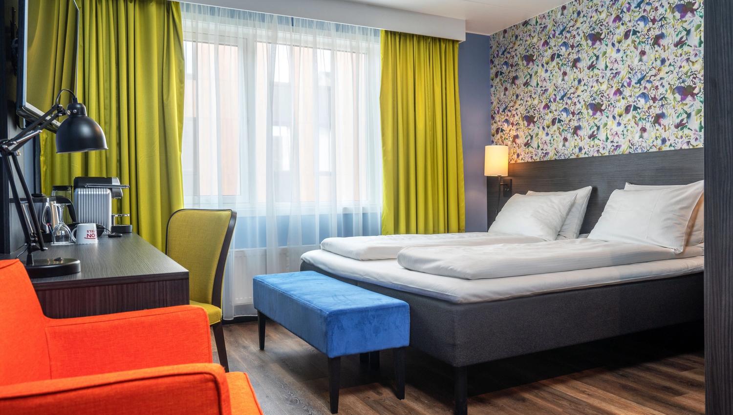 Modern hotel room with a double bed, colourful decor, and a large window.