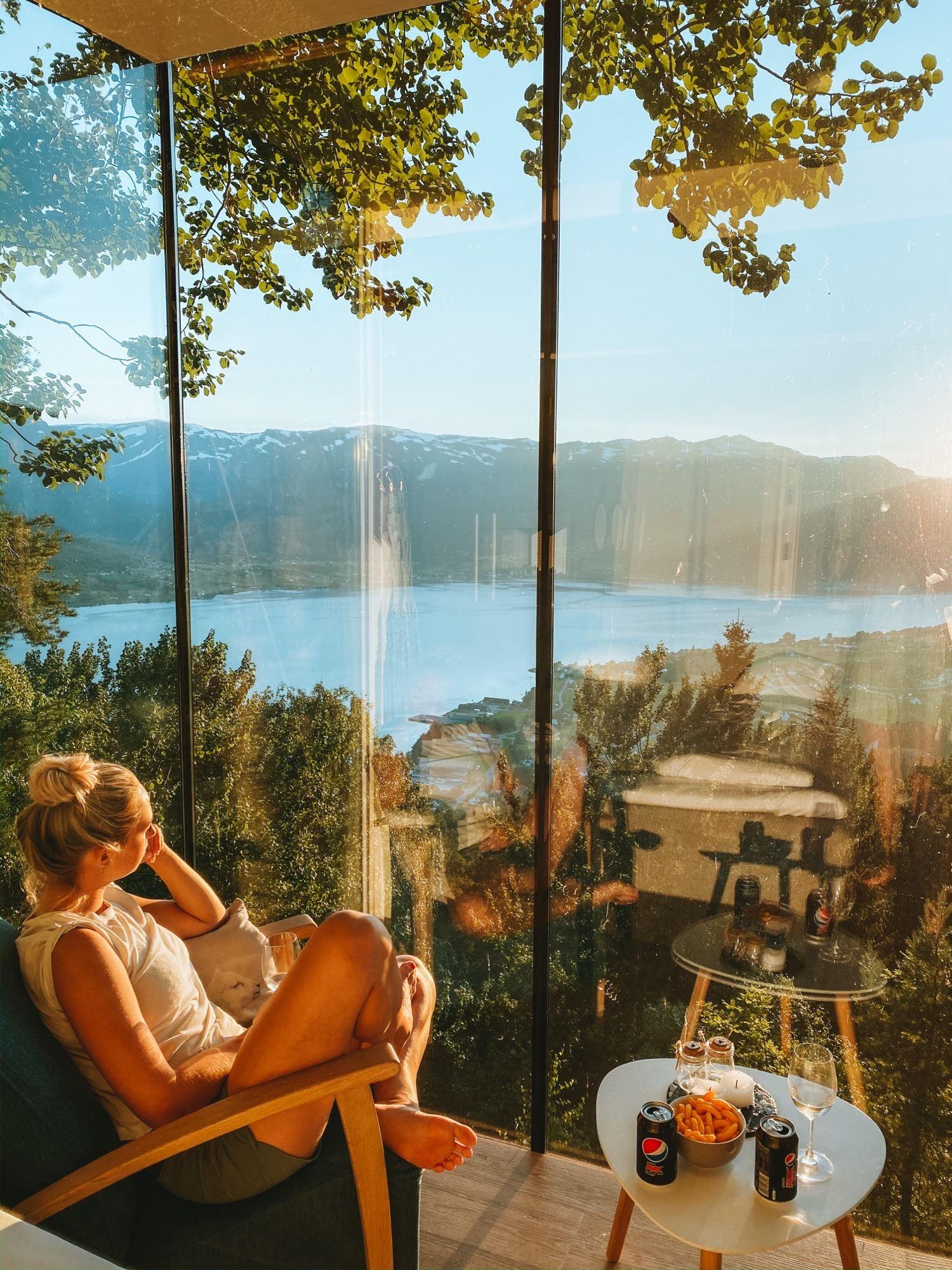 Person lounging in a chair by a large window, overlooking a sunlit lake, forested hills, and mountains..