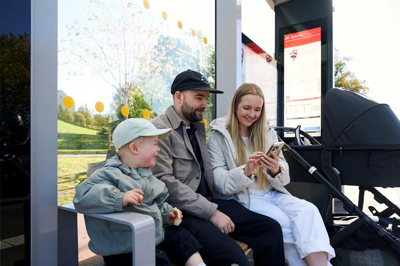 Mor, far og et barn som sitter på en benk ved et busstopp med en barnevogn og ser på en mobil.