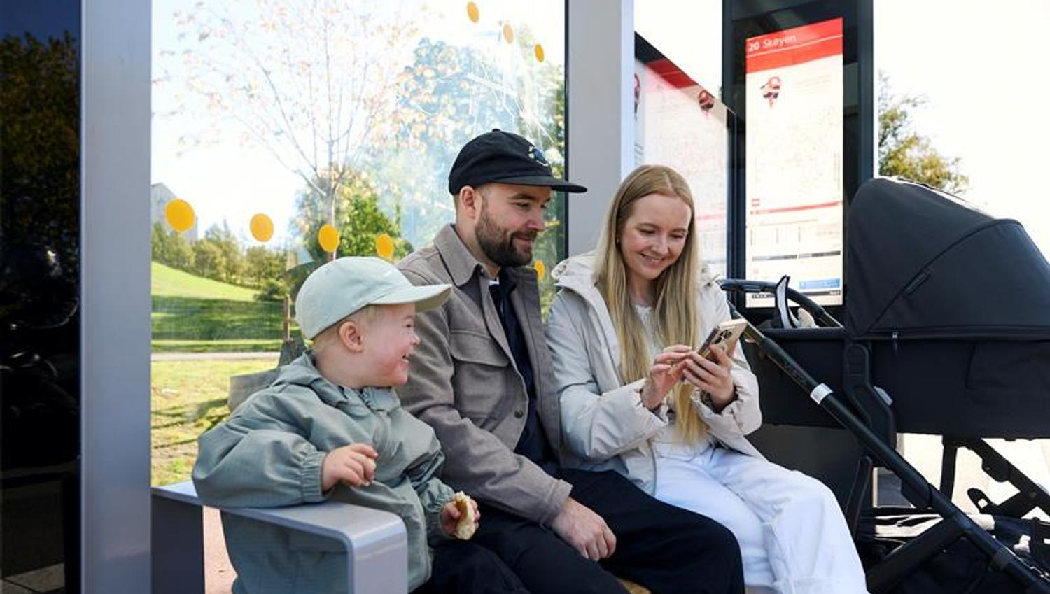 Mor, far og et barn som sitter på en benk ved et busstopp med en barnevogn og ser på en mobil.