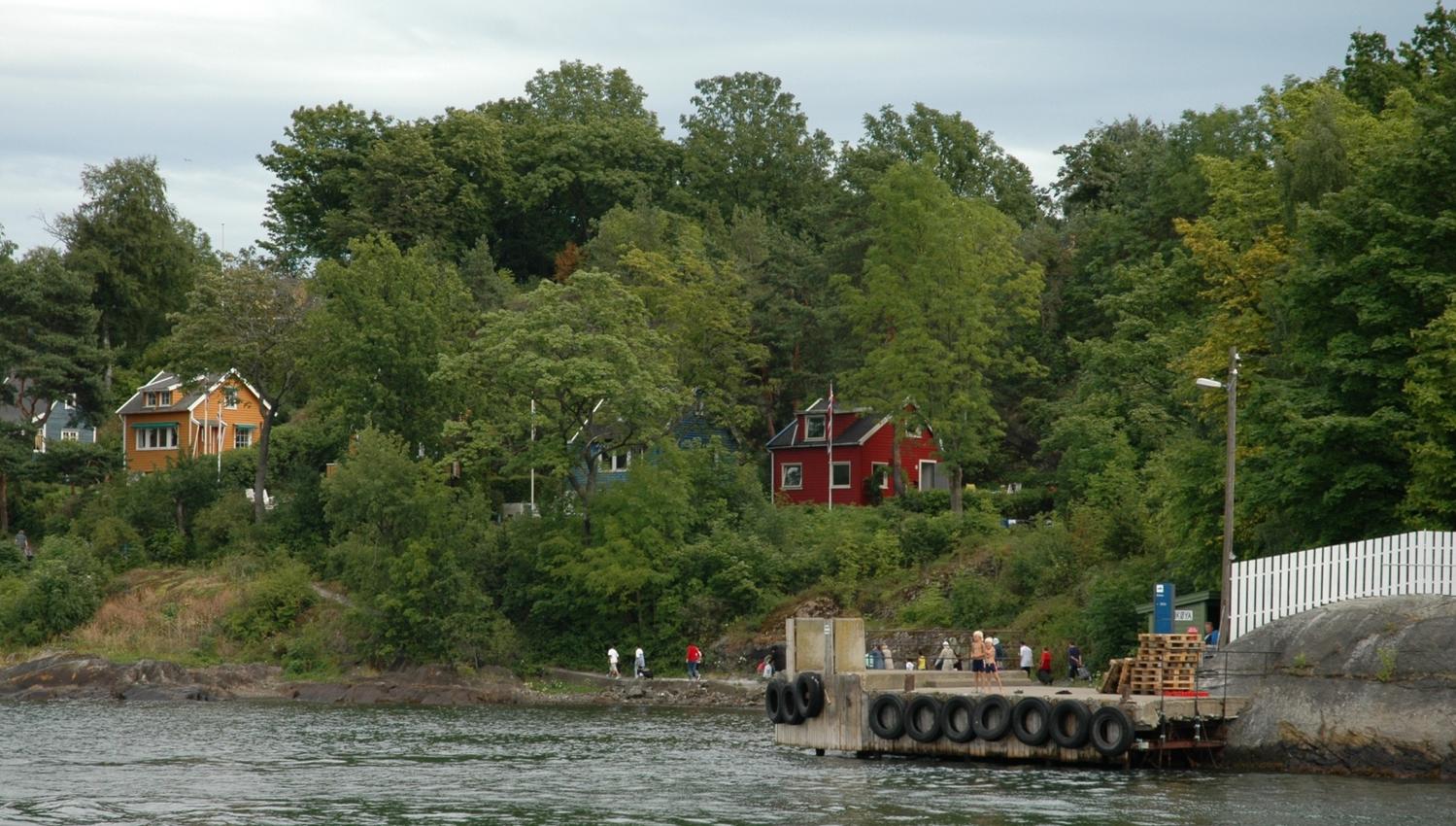 Brygga til øya med noen mennsker som går langs en sti, masse skog med noen trehus gjemt bak trærne.