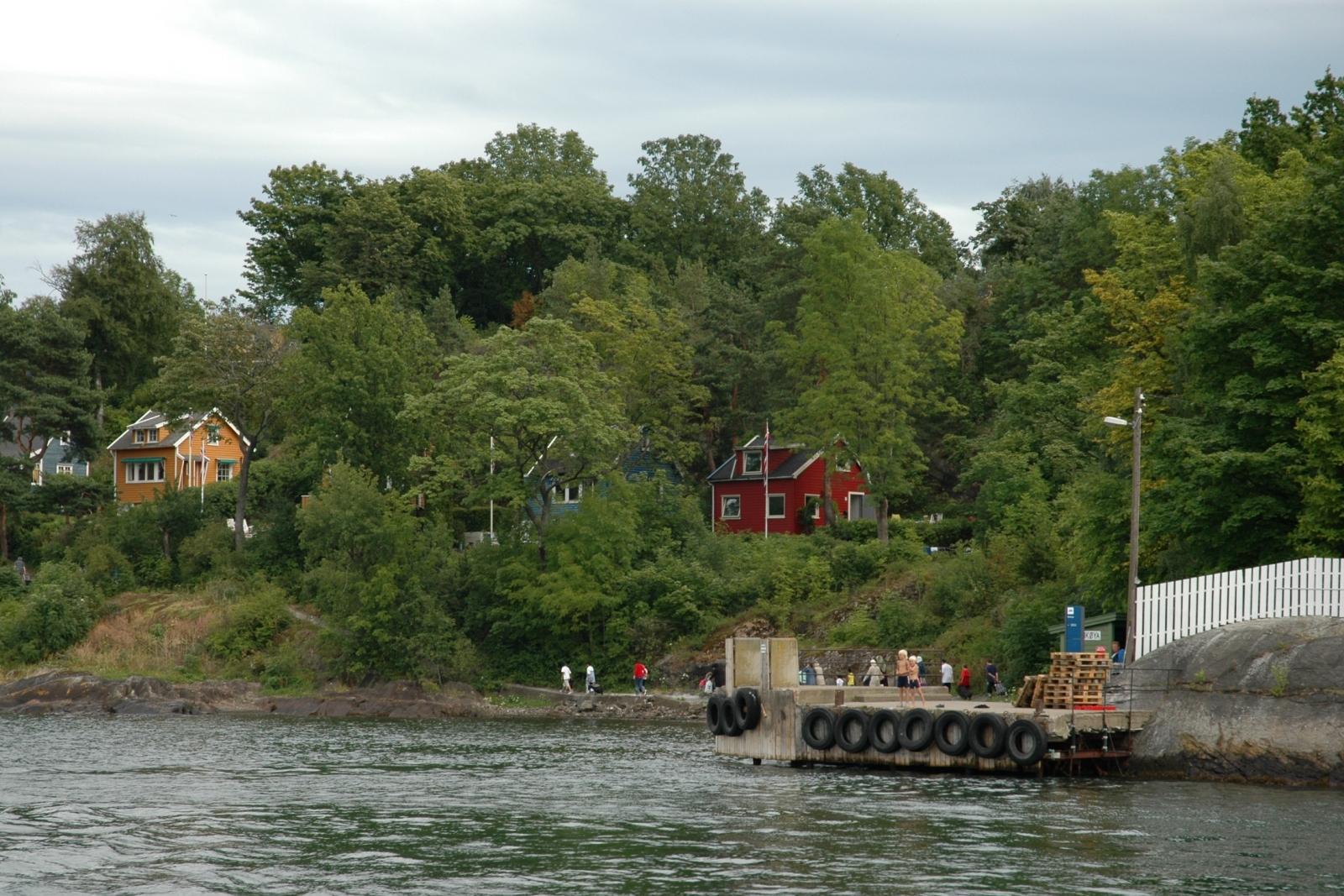 The pier to the island with a few people walking along a trail. A few wooden houses hidden in the trees.