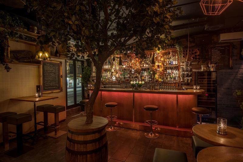 Bar interior with bar counter, bar stools, small tables and chairs, and a tree standing in a barrel in the middle of the room.