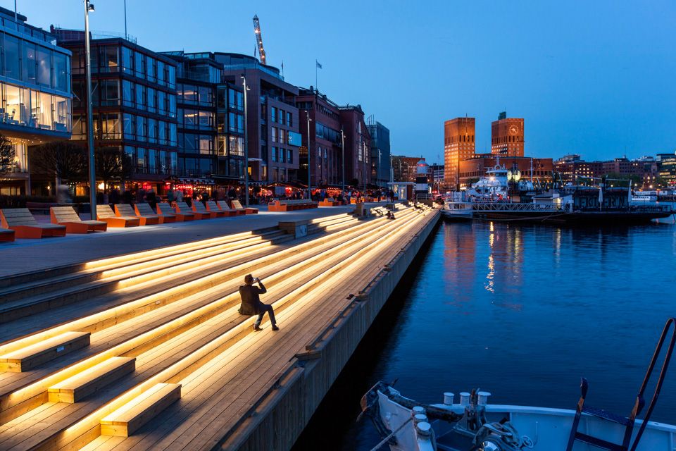Kveldsbilde av sitteseksjonen langs bryggen. Blå himmel. En siluett av en sittende person. Sitteseksjonen er opplyst av små lys. Rådhuset i bakgrunnen.