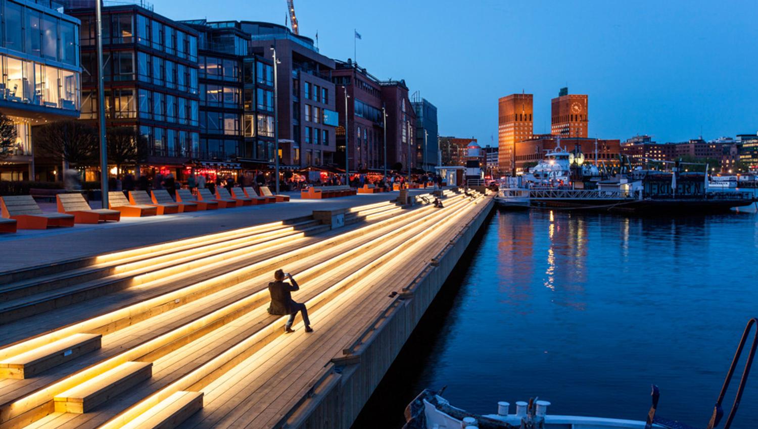 Kveldsbilde av sitteseksjonen langs bryggen. Blå himmel. En siluett av en sittende person. Sitteseksjonen er opplyst av små lys. Rådhuset i bakgrunnen.