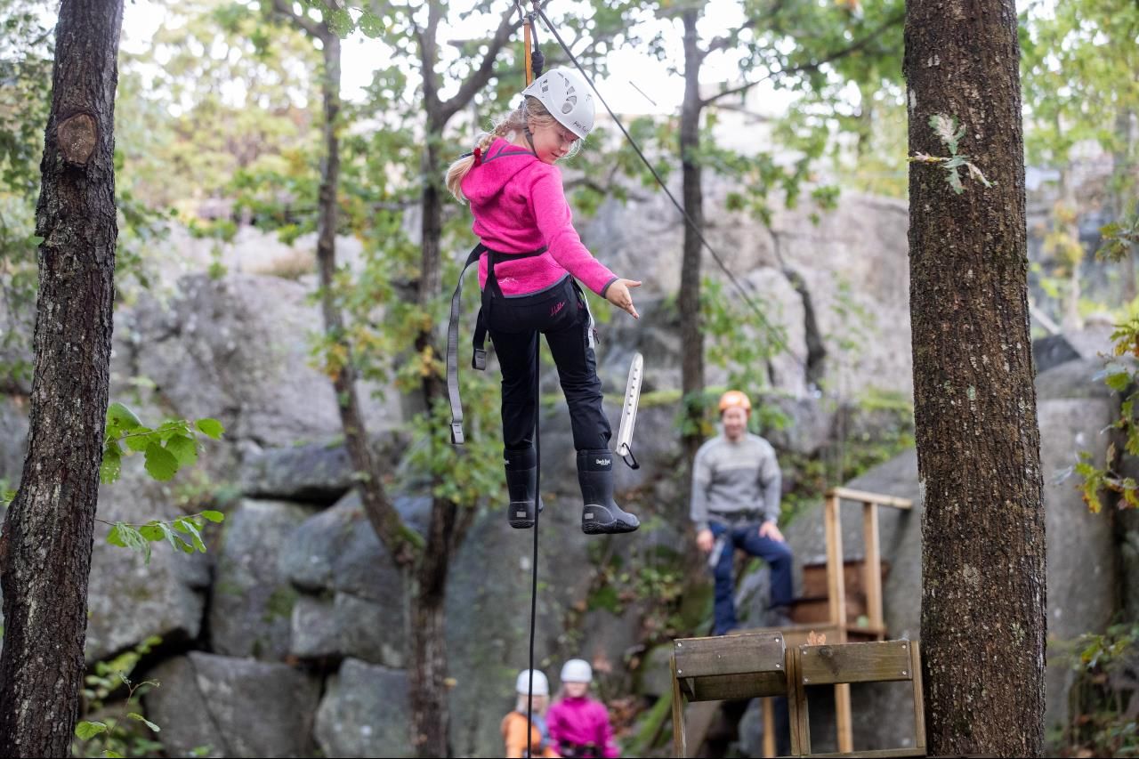 En ung jente henger fra zip line mellom trærne mens en voksen står klar til å ta henne imot