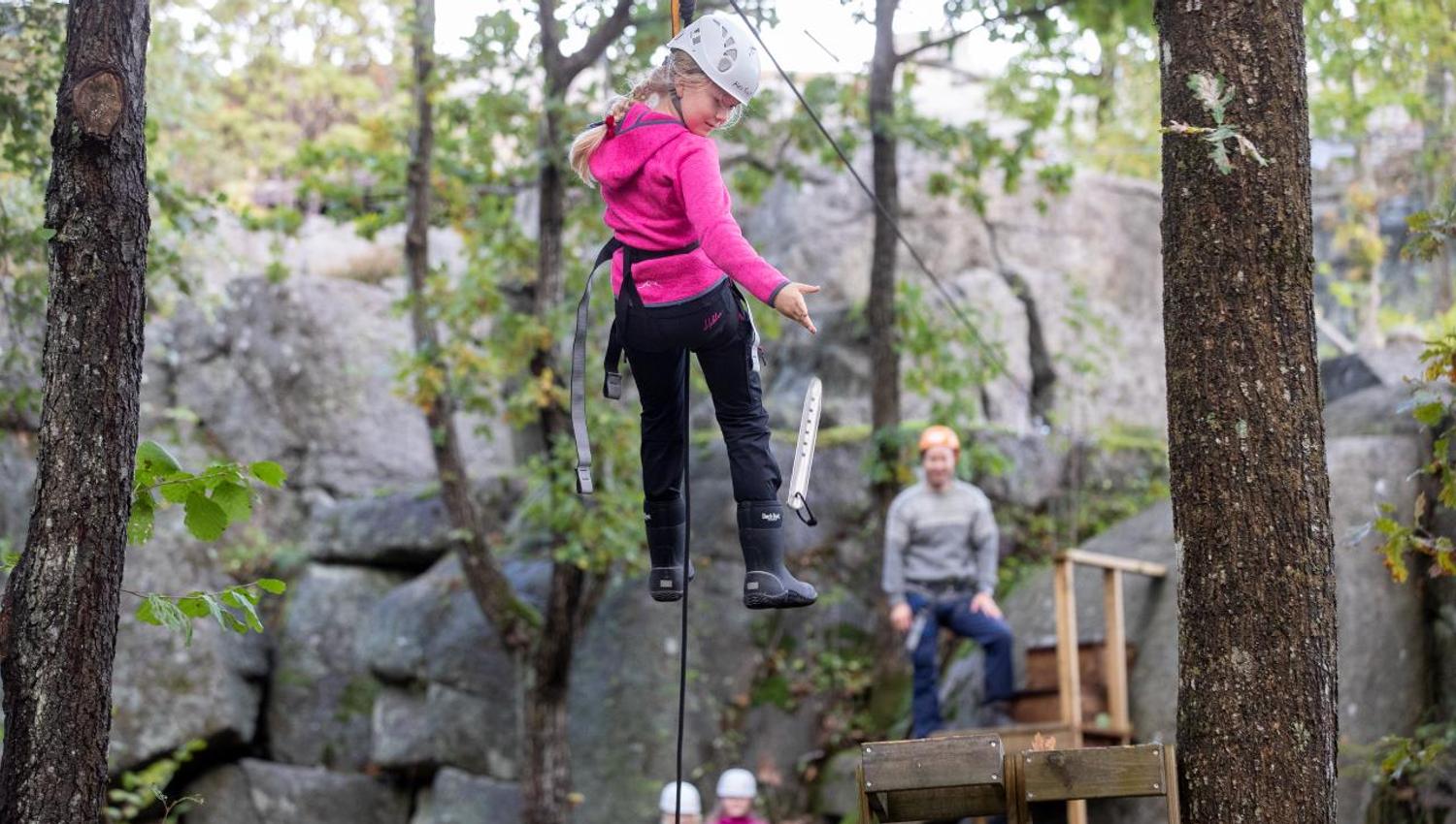 En ung jente henger fra zip line mellom trærne mens en voksen står klar til å ta henne imot