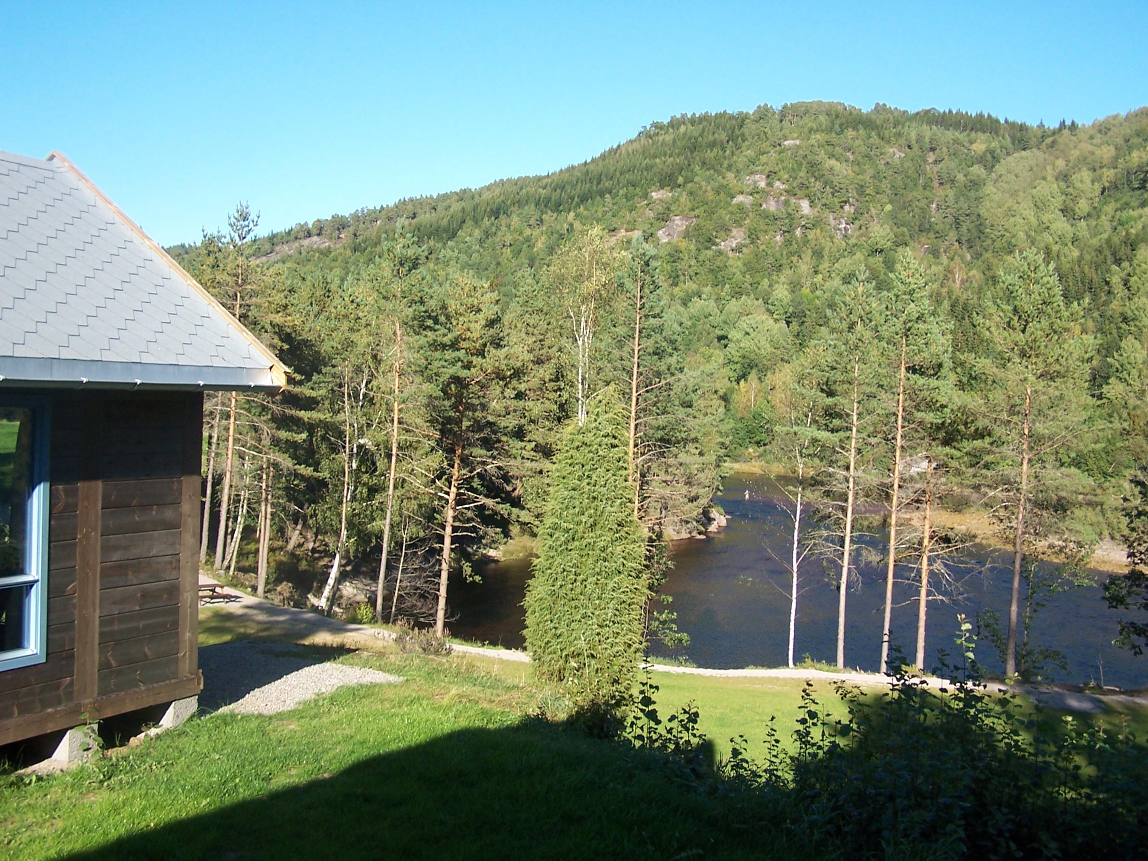 Brun laksehytte, grønn hage og vann nedenfor. Skog bak vannet, blå himmel.