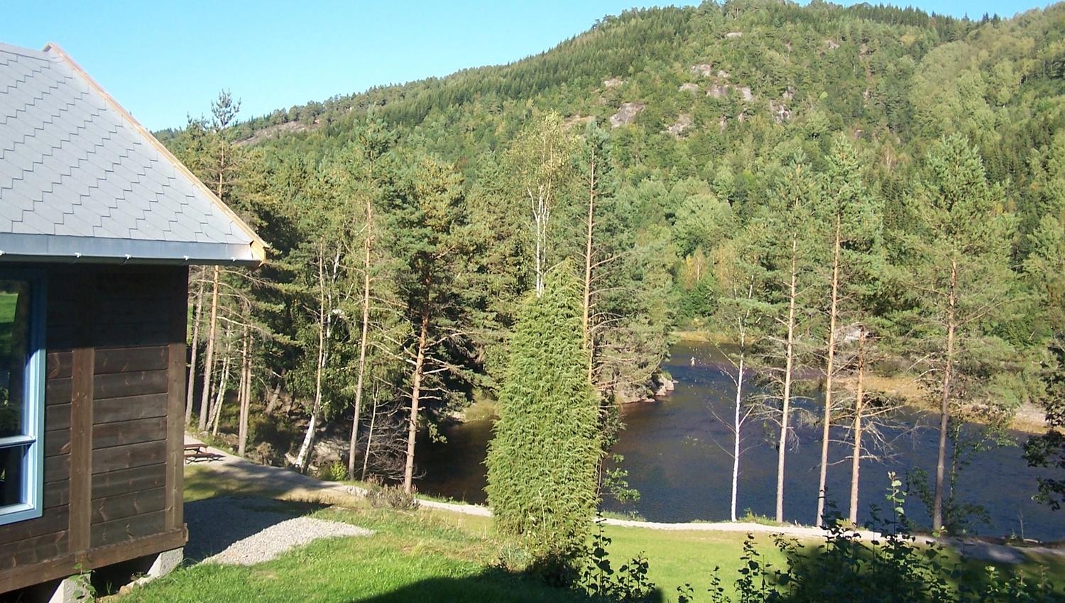 Brun laksehytte, grønn hage og vann nedenfor. Skog bak vannet, blå himmel.