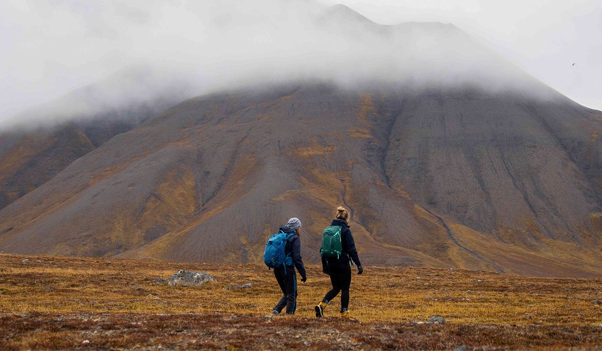 Two guests hiking on the tundra with clouds and a mountain in the background