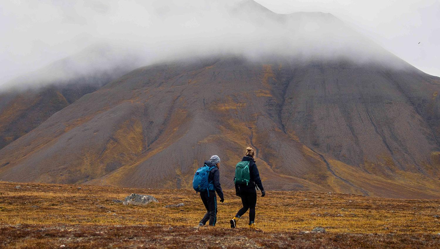 Two guests hiking on the tundra with clouds and a mountain in the background