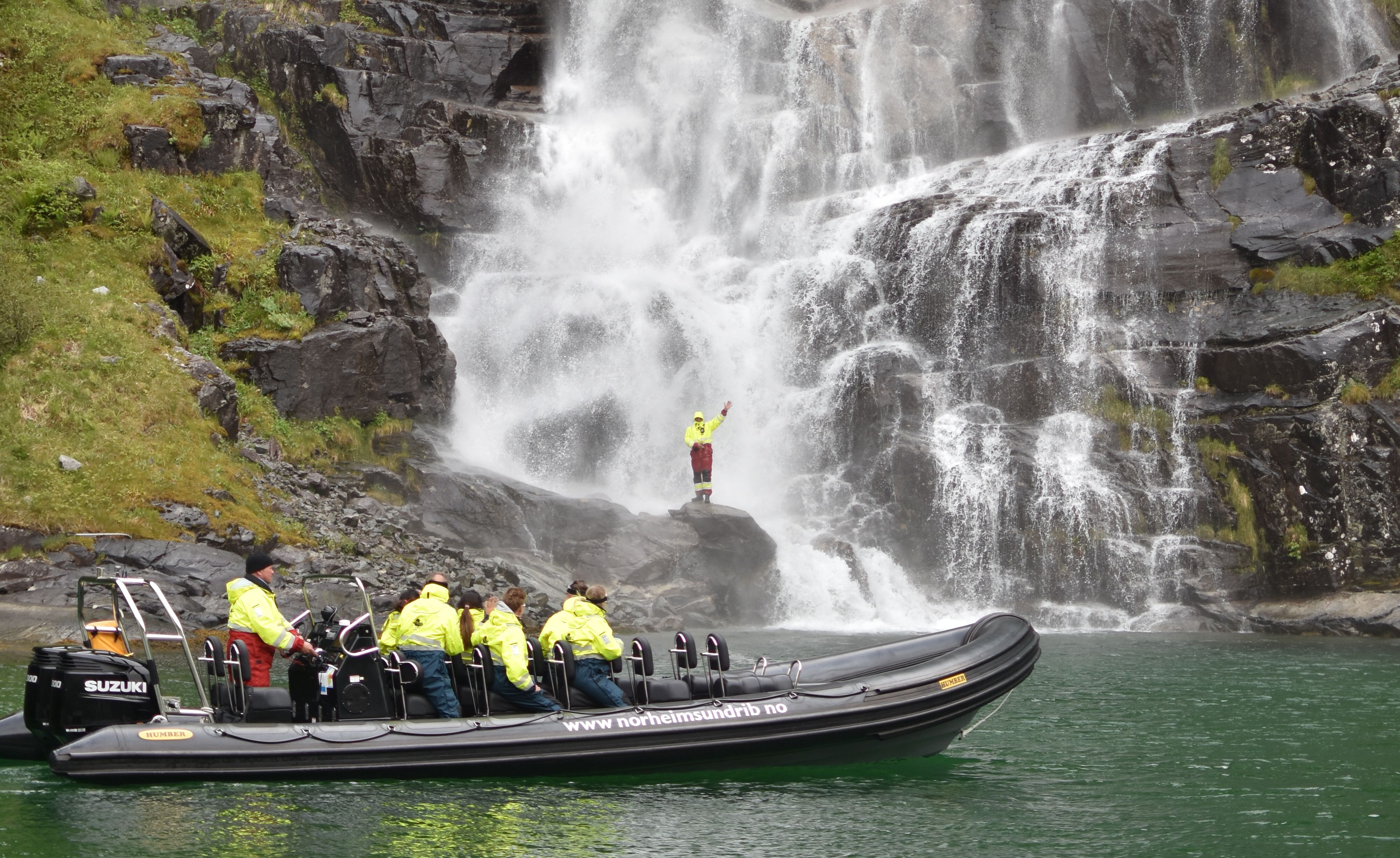 Ein eventyrlysten deltakar står på ein stein nær fossen medan resten av gruppa ser på frå RIB-båten.