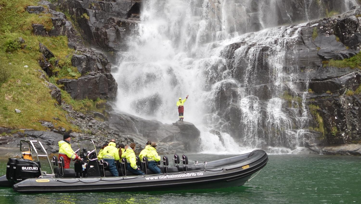 Ein eventyrlysten deltakar står på ein stein nær fossen medan resten av gruppa ser på frå RIB-båten.