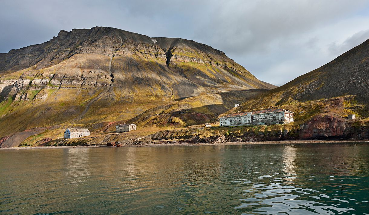 Abandoned buildings in the former mining community Grumant along the shores of the fjord Isfjorden in the foreground, with tall mountains and a cloudy