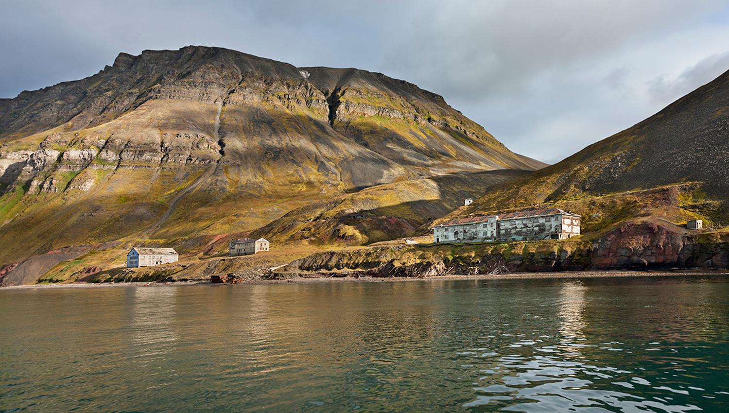 Abandoned buildings in the former mining community Grumant along the shores of the fjord Isfjorden in the foreground, with tall mountains and a cloudy