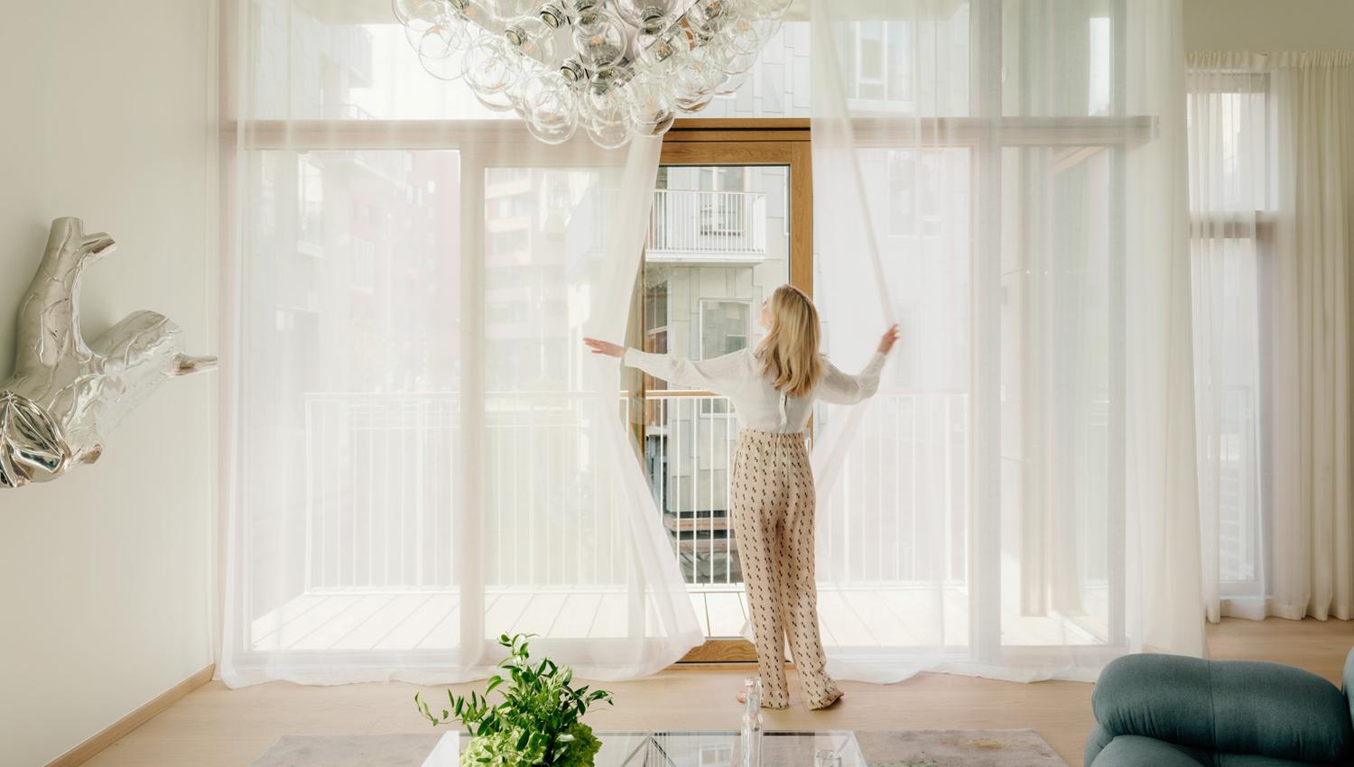Lady in white living room with large panoramic windows pulling the curtains aside