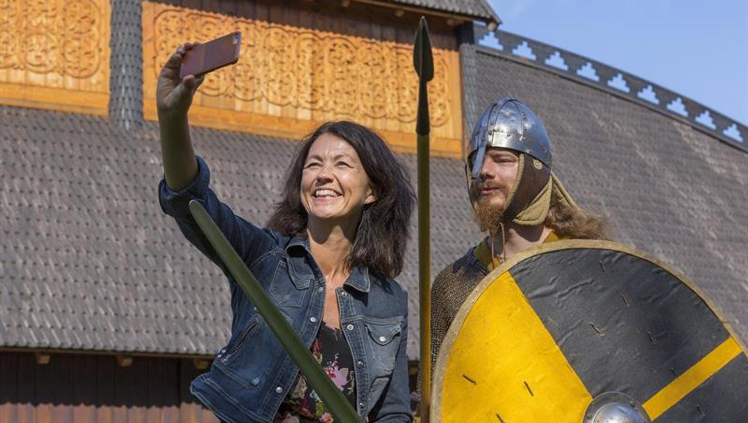 En dame og en mann kledd ut som en viking står foran Gildehallen og tar en selfie.