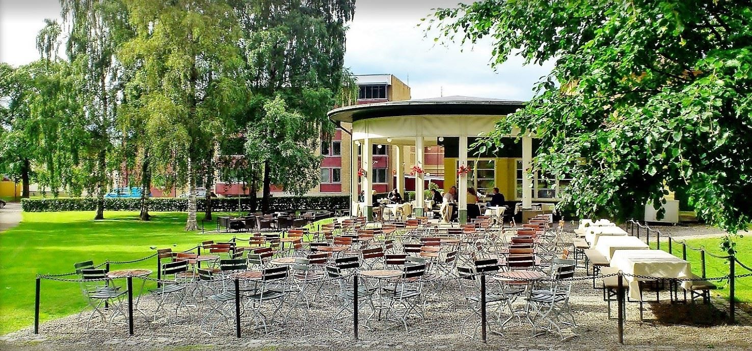 Mange tomme metallstoler og bord står på grus foran en rund kafépaviljong i en grønn park med trær..