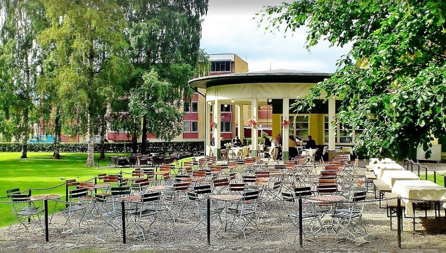Mange tomme metallstoler og bord står på grus foran en rund kafépaviljong i en grønn park med trær..