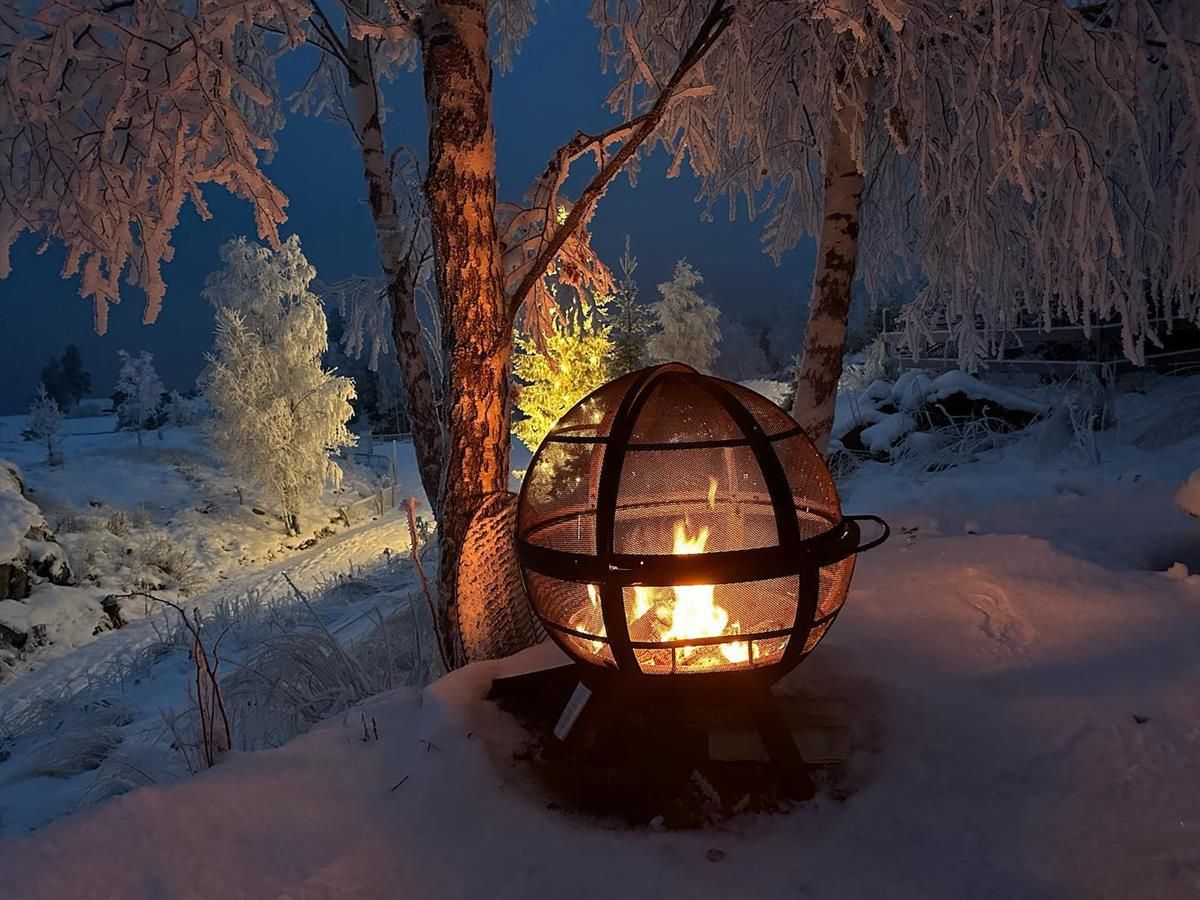 En lykt med åpen flamme står i snødekt vinterlandskap under trær i skumring.