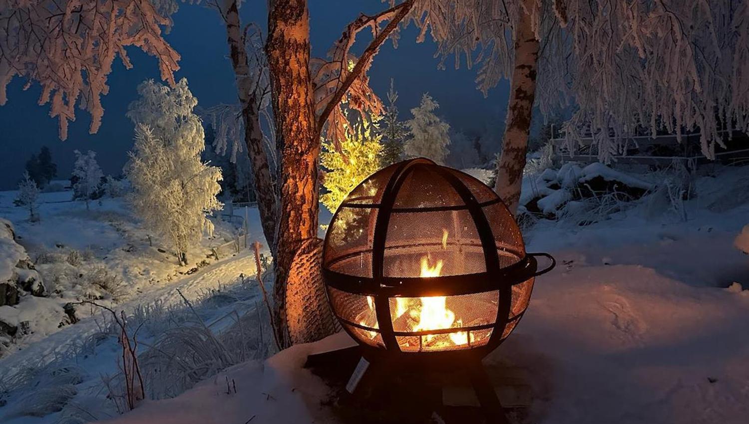 En lykt med åpen flamme står i snødekt vinterlandskap under trær i skumring.