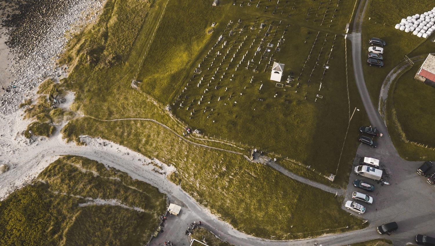 Luftfoto av ei kyrkjegard på grønt jorde ved ei steinete strand, med veg, parkerte bilar og nokre bygg..