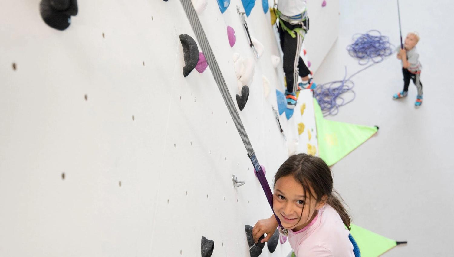Two children climbing an indoor wall while a third person belays from the ground.