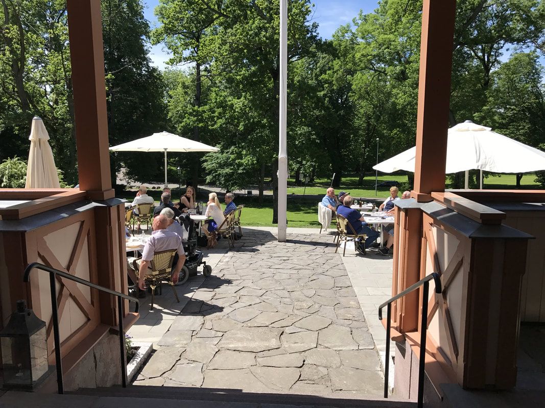 Photo from the outdoor dining area. Platting with slate stone and tables with parasols. There are people sitting at almost all the tables and the sun 