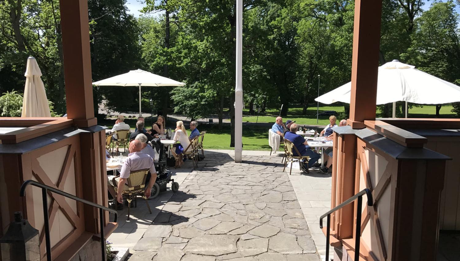 Photo from the outdoor dining area. Platting with slate stone and tables with parasols. There are people sitting at almost all the tables and the sun