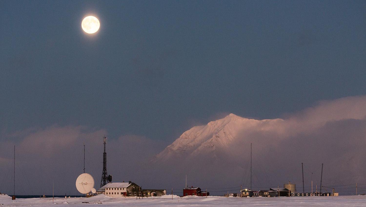 Isfjord Radio in a snowy landscape with mist over the fjord in the background and the moon shining above.