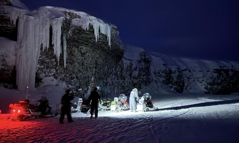 A group of people are standing and walking around some snowmobiles in the dark rigtht below a cliff with frozen ice hanging down from it.