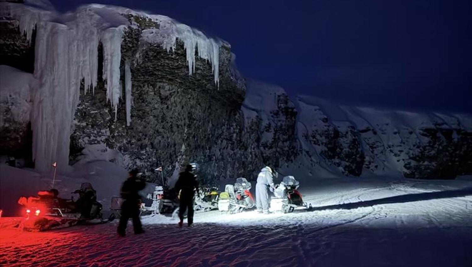 A group of people are standing and walking around some snowmobiles in the dark rigtht below a cliff with frozen ice hanging down from it.