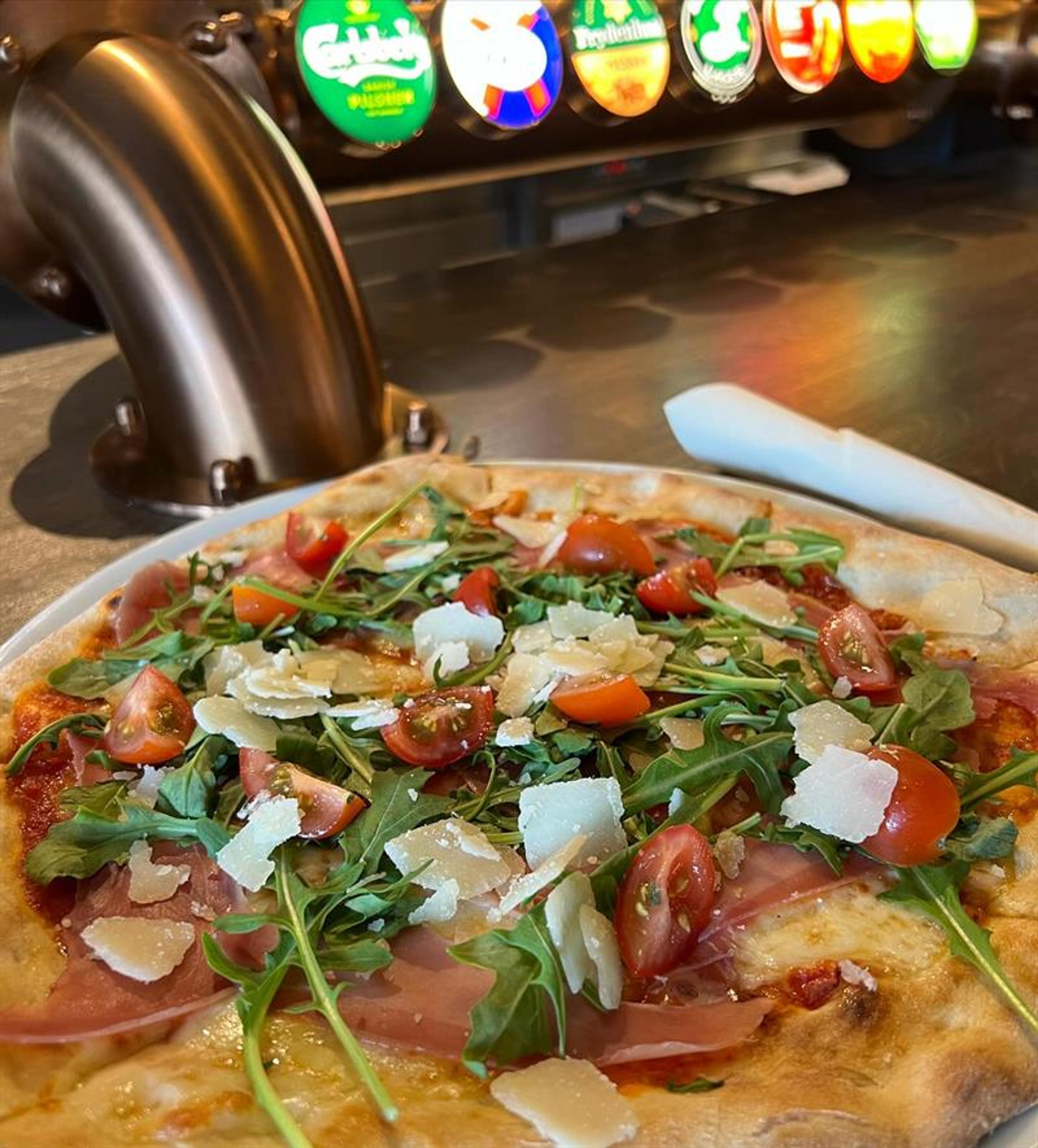 Nærbilde av en pizza toppet med ferske grønnsaker, urter og smeltet ost. Fargerike brusdispensere er synlige i bakgrunnen.