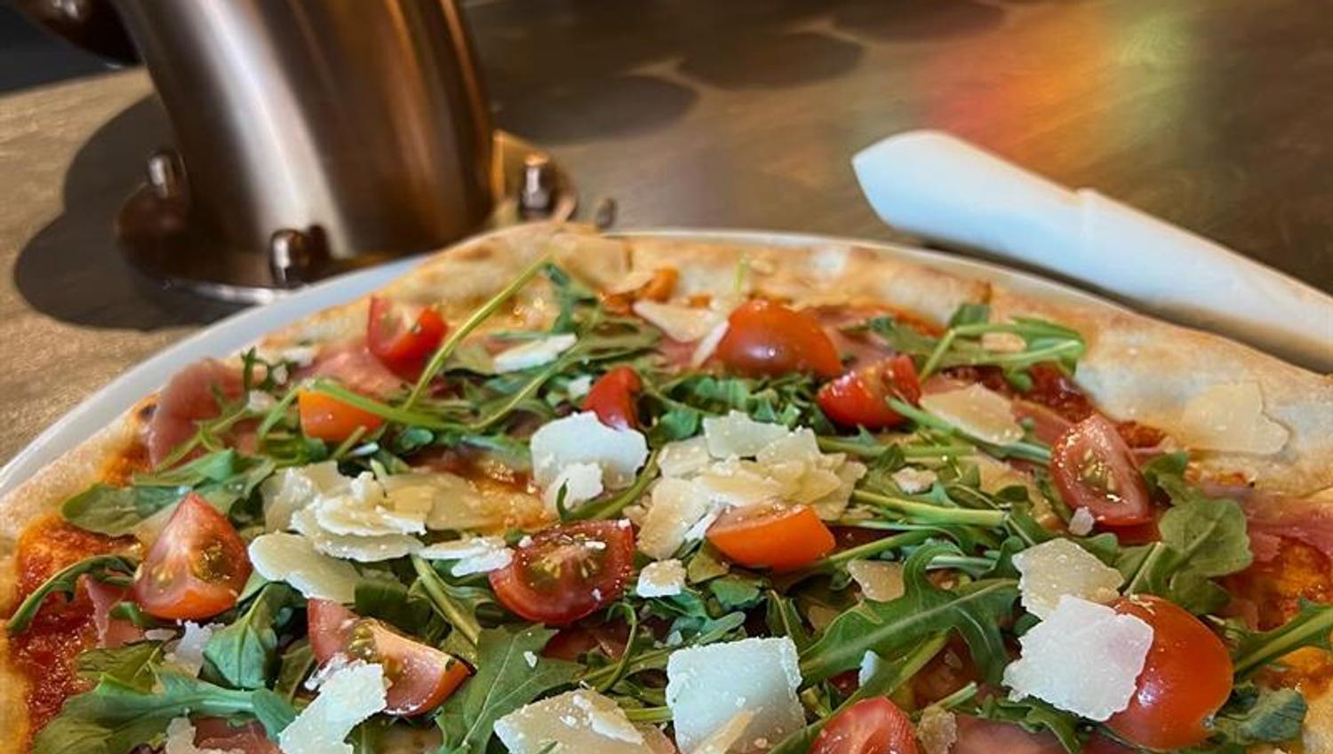 Nærbilde av en pizza toppet med ferske grønnsaker, urter og smeltet ost. Fargerike brusdispensere er synlige i bakgrunnen.