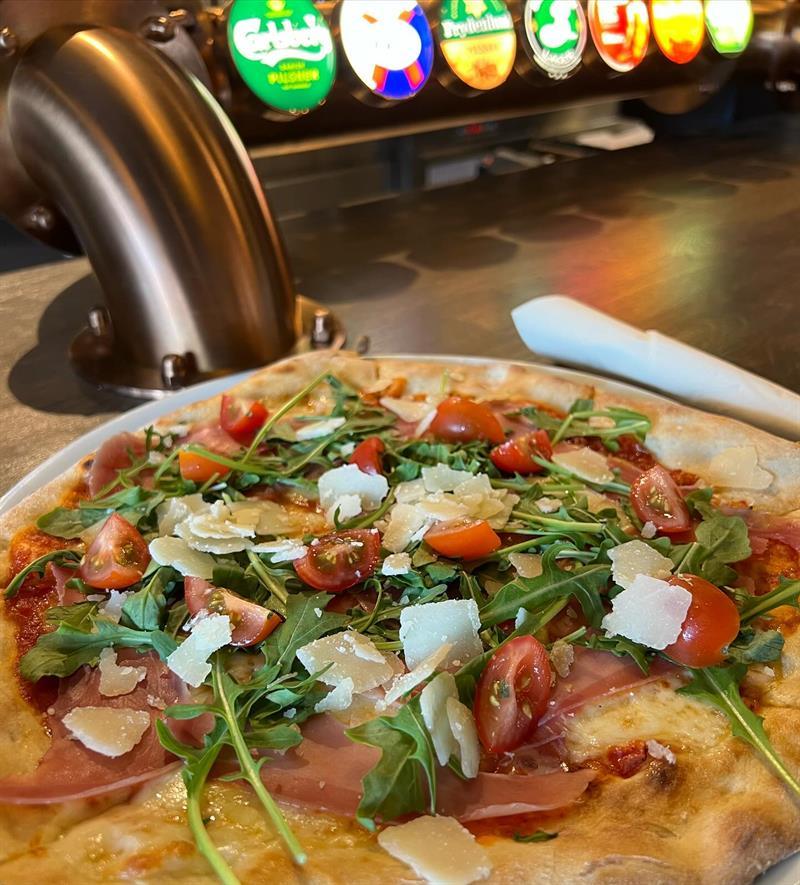 Nærbilde av en pizza toppet med ferske grønnsaker, urter og smeltet ost. Fargerike brusdispensere er synlige i bakgrunnen.