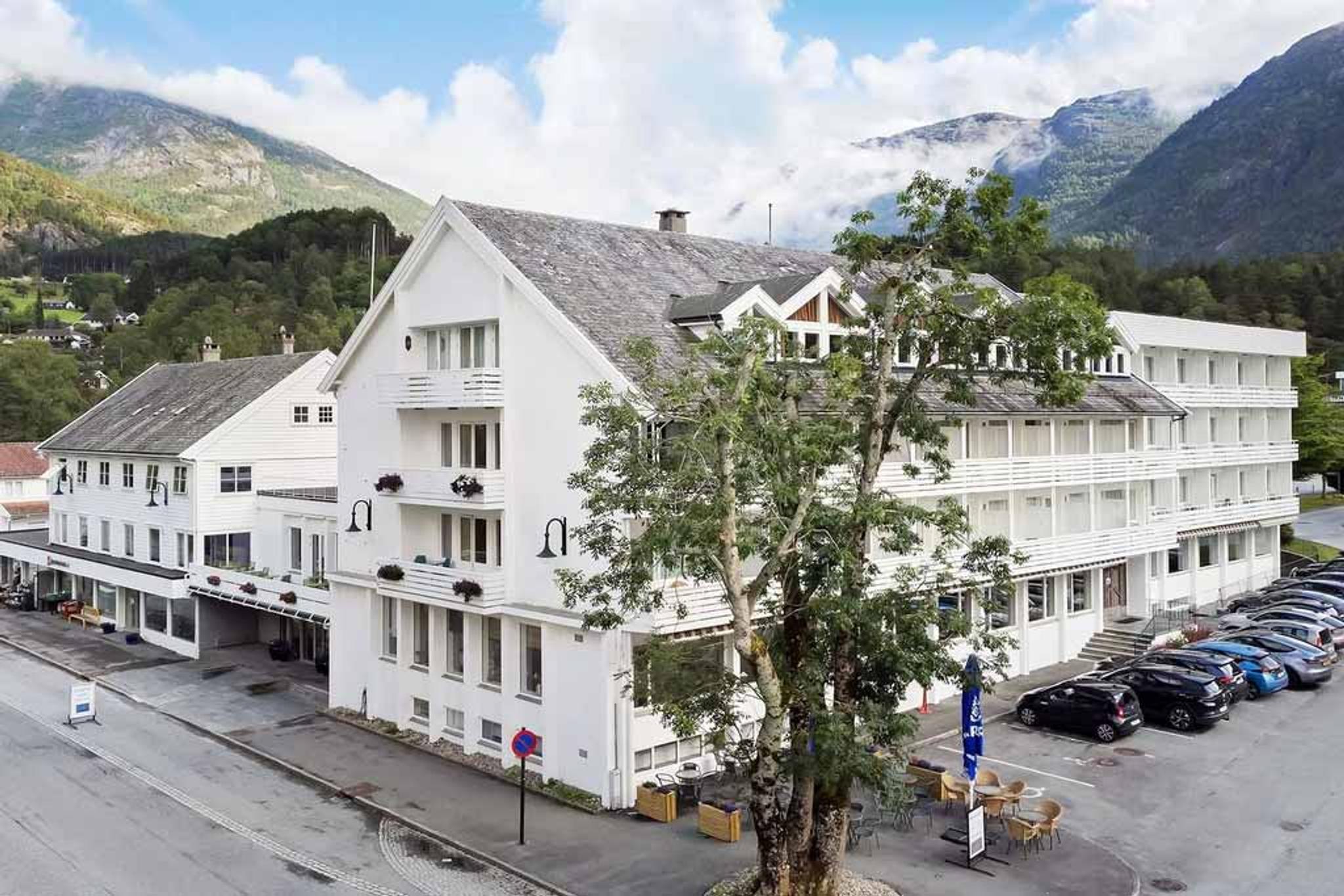 Fasaden til Kinsarvik Fjordhotel med idyllisk utsikt mot Hardangerfjorden, omgitt av vakker natur og fjellandskap.