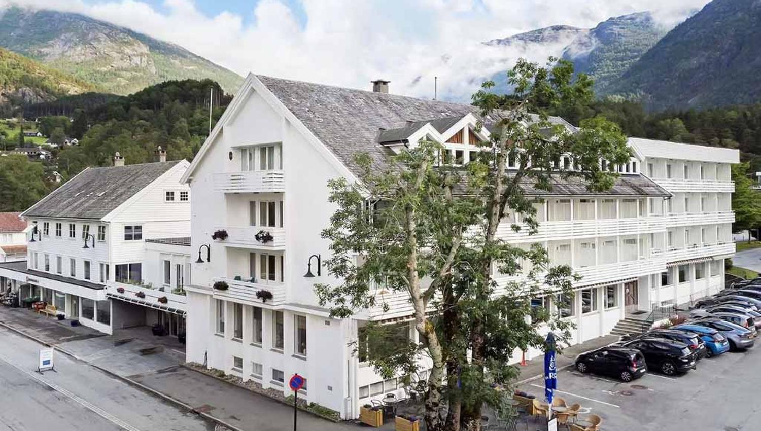 Fasaden til Kinsarvik Fjordhotel med idyllisk utsikt mot Hardangerfjorden, omgitt av vakker natur og fjellandskap.