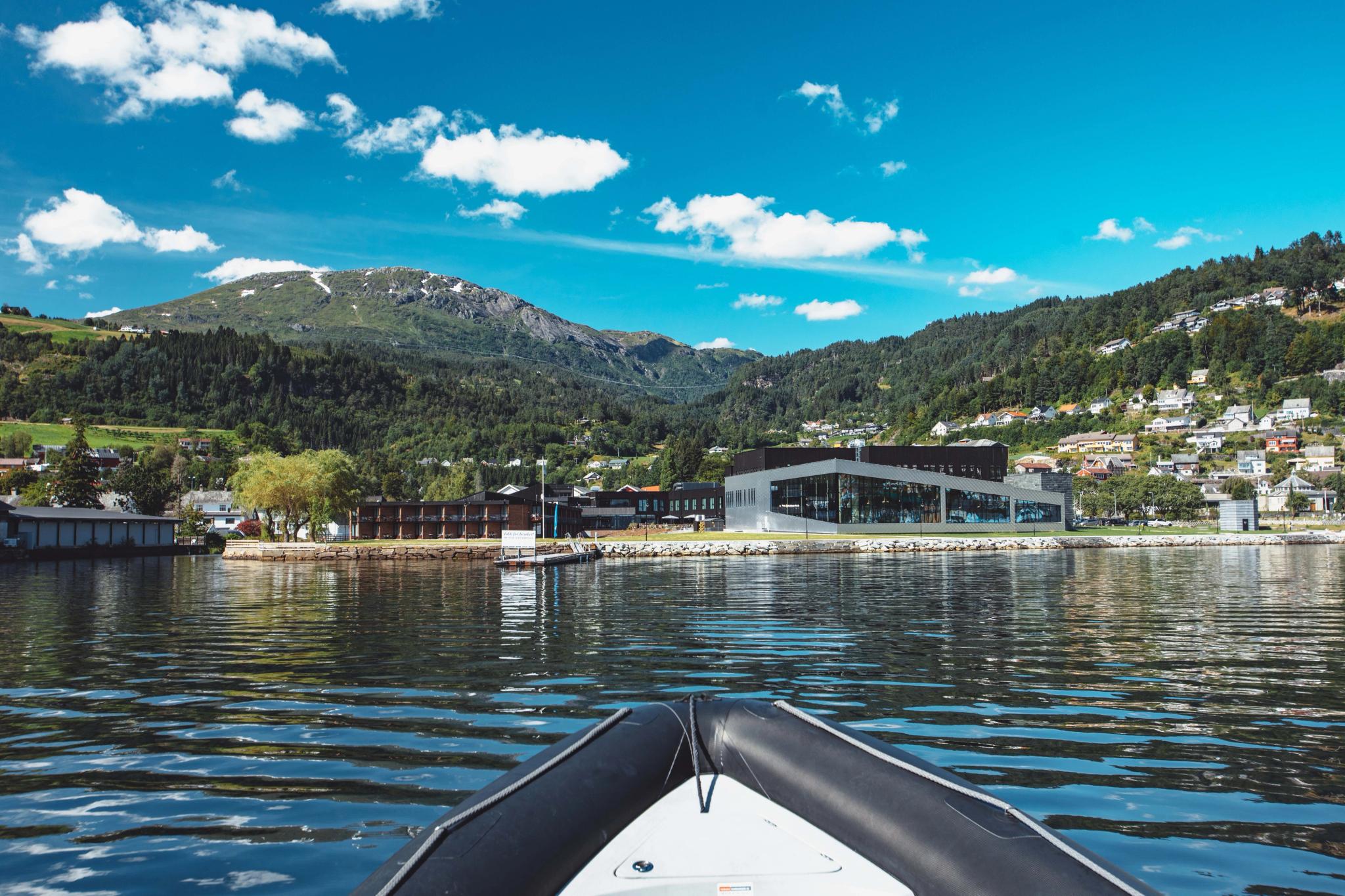 View of Hardangerfjord Hotel from the fjord, surrounded by mountains and a charming village.