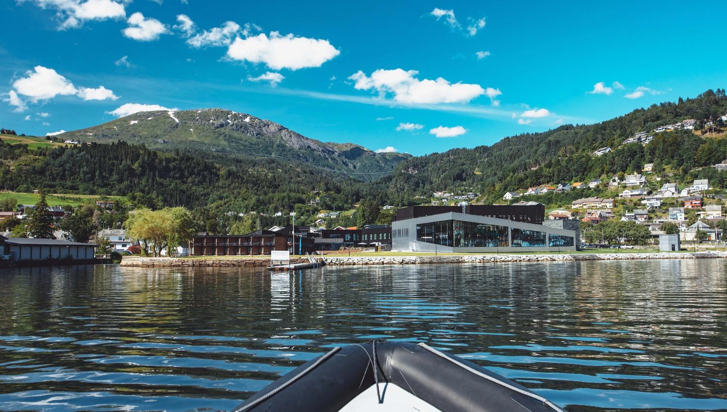 Utsikt mot Hardangerfjord Hotel frå fjorden, med fjell og bygd i bakgrunnen.