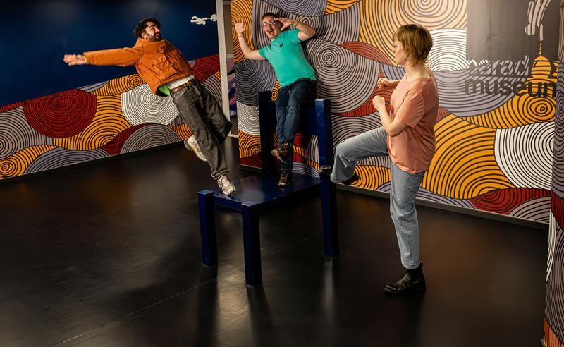 Tre personer poserer lekent i en interaktiv museumsinstallasjon som skaper en illusjon av at de mister balansen.