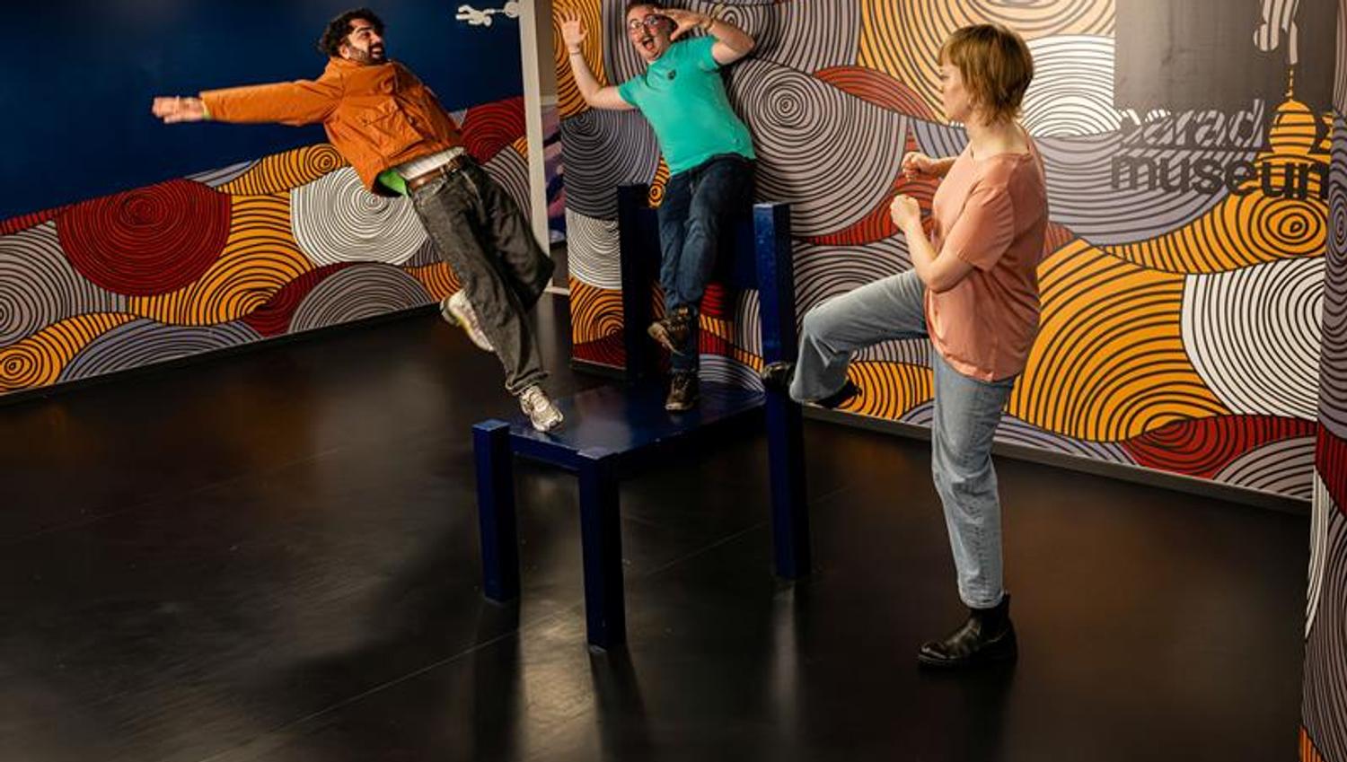 Tre personer poserer lekent i en interaktiv museumsinstallasjon som skaper en illusjon av at de mister balansen.
