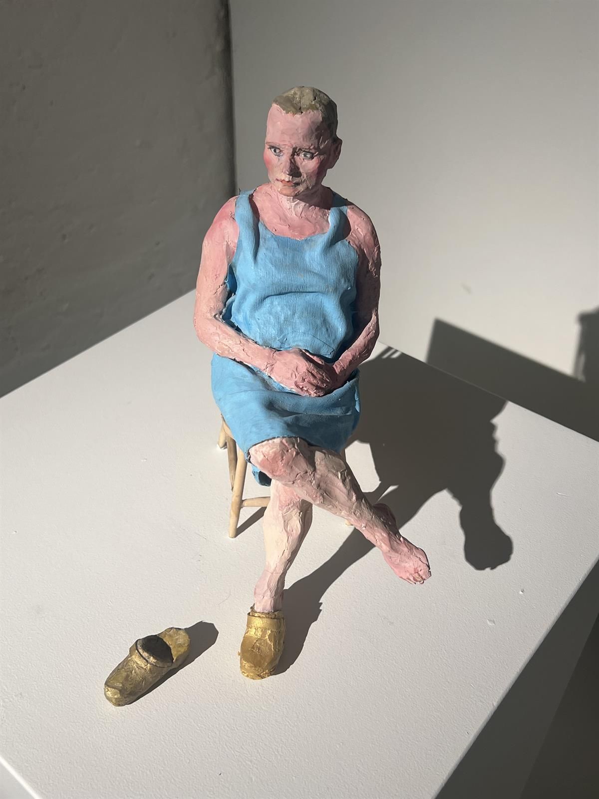 Skulptur av en sittende figur i blå kjole med sko plassert på gulvet foran.