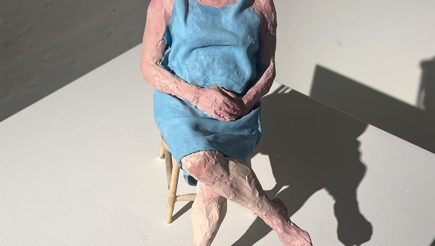 Skulptur av en sittende figur i blå kjole med sko plassert på gulvet foran.