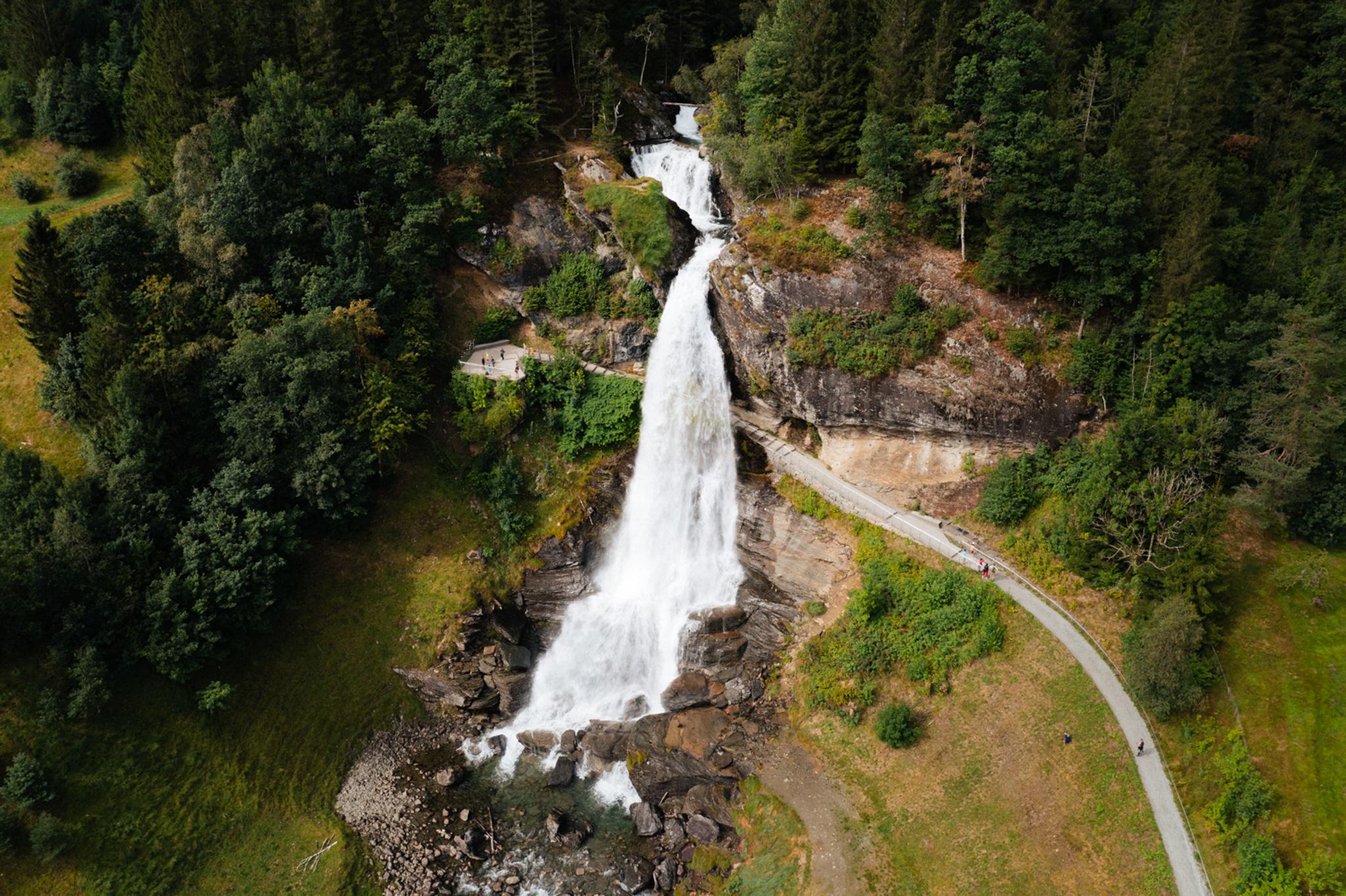 Aerial view of Steinsdalsfossen, a majestic waterfall surrounded by lush greenery and a walking path, with people strolling close to the falls in Norw